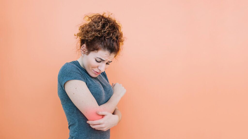 A woman grimaces in pain, clutching her elbow against a peach backdrop, a common ache pains treatment in Chicago urgent care clinics. This image illustrates the everyday pains managed in local healthcare settings.