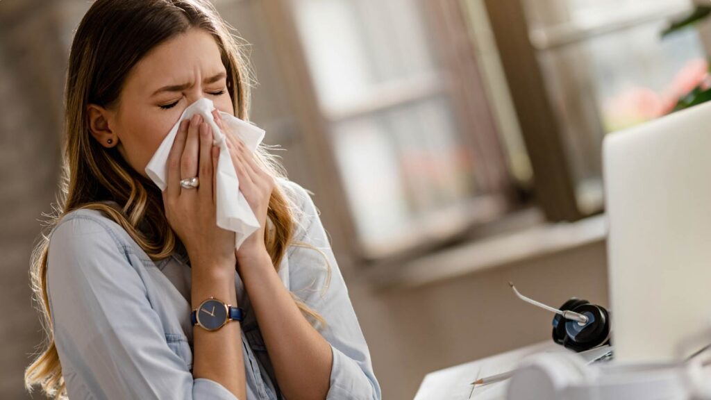 A woman is pictured blowing her nose into a tissue, which suggests she may be seeking sinus treatment in Illinois urgent care. The image captures a moment of discomfort that could indicate common symptoms addressed at an urgent care clinic.